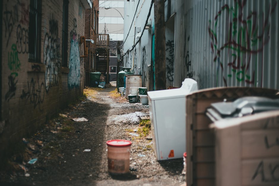 A narrow, outdoor back alleyway between two brick and corrugated metal buildings, with the side of one building covered in graffiti. The alley is cluttered with various debris and discarded items, including plastic buckets, a washing machine, or dryer, and other miscellaneous waste containers. The ground is uneven, muddy, and scattered with small patches of moss or grass, indicating a lack of maintenance. Several large, dark green wheelie bins are lined up along the brick wall, suggesting use for waste collection or recycling. In the background, a staircase leading to an upper floor or rooftop is visible, along with faint indications of other urban waste and rubbish accumulation. The lighting appears natural, with diffuse daylight illuminating the scene, emphasizing the gritty, neglected environment typical of an area requiring rubbish removal or private waste disposal services. Waste Clearance Lambeth may handle such privately handled waste in urban backstreets, supporting alternative waste clearance solutions outside municipal rubbish collection. 