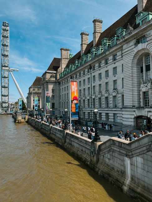 A wide view of a historic white sandstone building with multiple chimneys and green rooftop dormer windows, situated along the edge of a river. The building features arched windows and detailed architectural elements. In front of the building, a paved promenade runs parallel to the river, with a crowd of pedestrians walking and standing along the railings. Several tall light posts line the walkway, and banners featuring characters from a popular video game are mounted on lampposts close to the building. To the left, a modern tower with a narrow, vertical structure and a crane are visible in the background, while the river’s water appears muddy and surrounds the promenade area. The sky is partly cloudy with blue visible, creating a bright and clear atmosphere. This scene exemplifies urban riverside environments where private waste removal services might operate in conjunction with public areas, highlighting the importance of effective rubbish disposal for busy metropolitan settings like Lambeth.
