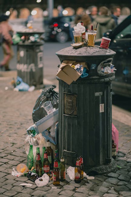 A black metal rubbish bin with a domed lid is positioned on a brick-paved street, surrounded by overflowing litter including several glass bottles, some beer bottles with green, brown, and clear glass, and crumpled paper scattered on the ground. Articles such as a cardboard box and plastic packaging are lodged in the side of the bin. The top of the bin holds discarded items resembling food wrappers, paper cups with lids, and glasses containing drinks, some with straws, indicating recent use. In the background, there is a busy street scene with parked cars, a grey trash bin, and blurred figures of pedestrians, suggesting an urban environment with high foot traffic. The lighting appears natural, possibly from outdoor daylight, emphasizing the mess around the bin. This scene exemplifies improper waste disposal and highlights the importance of scheduled rubbish collection services like those provided by Waste Clearance Lambeth for maintaining cleanliness and managing waste effectively in urban areas.