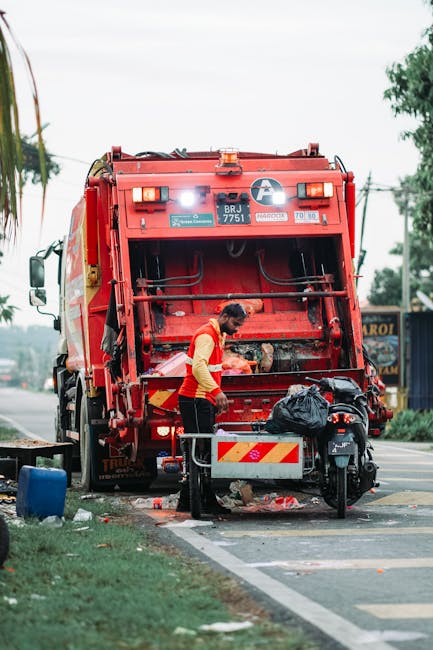 A waste collection worker wearing an orange high-visibility vest is operating a large red garbage truck, which is actively engaged in waste collection on a street. The back of the truck is open, revealing mechanical components and a partly visible hopper for waste intake. The worker is standing close to a black motorbike that is parked alongside the truck, with a black plastic bag placed on top of the motorcycle. The truck is positioned on the side of a paved road, with a curb separating it from a grassy verge and sidewalk. In the background, there are trees, utility poles with wires, and a roadside signboard partly obscured by the truck. The environment appears to be during daylight hours, with overcast skies providing diffused natural light. The scene captures an on-site waste collection activity, which may be an example of private or independent rubbish removal, illustrating the type of equipment and personnel involved in rubbish services like those provided by Waste Clearance Lambeth.
