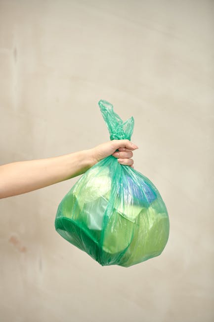 A person's arm and hand are visible, holding a green and blue plastic rubbish bag by its twisted top. The bag appears to contain general waste and is slightly translucent, revealing the contents inside. The background consists of a plain, neutral beige wall, with no other objects or environmental features visible. The scene suggests an act of waste disposal, likely part of household or commercial rubbish removal, which aligns with services offered by Waste Clearance Lambeth. The lighting is soft and even, without harsh shadows or reflections, emphasizing the bag's texture and color. The image reflects a typical moment of private waste handling before collection or transfer to a disposal facility, illustrating the importance of proper rubbish management in the context of independent clearance or non-local-authority waste disposal methods.