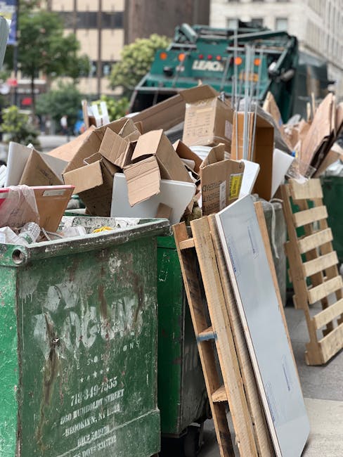 A waste collection worker wearing an orange high-visibility vest is operating a large red garbage truck, which is actively engaged in waste collection on a street. The back of the truck is open, revealing mechanical components and a partly visible hopper for waste intake. The worker is standing close to a black motorbike that is parked alongside the truck, with a black plastic bag placed on top of the motorcycle. The truck is positioned on the side of a paved road, with a curb separating it from a grassy verge and sidewalk. In the background, there are trees, utility poles with wires, and a roadside signboard partly obscured by the truck. The environment appears to be during daylight hours, with overcast skies providing diffused natural light. The scene captures an on-site waste collection activity, which may be an example of private or independent rubbish removal, illustrating the type of equipment and personnel involved in rubbish services like those provided by Waste Clearance Lambeth.