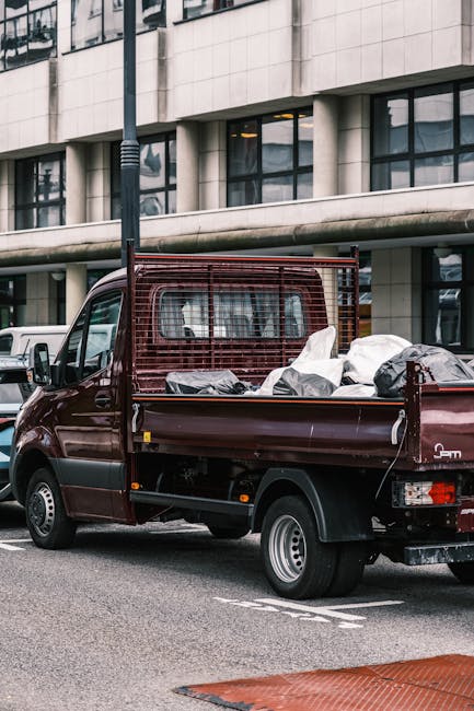 A small burgundy flatbed truck parked on a city street in front of a modern office building with large glass windows and white exterior panels. The truck's cargo area is filled with various discarded items, including bags and packaging, indicating it is used for rubbish removal. The truck features a protective metal grid at the cab's rear window to secure the load. Surrounding the vehicle are other parked cars, a pavement, and a street lamp, with muted lighting suggesting overcast weather. The scene reflects a typical urban environment where private waste disposal services, such as those provided by Waste Clearance Lambeth, handle alternative rubbish collection outside of municipal systems, focusing on on-site clearance and private waste handling solutions. The overall atmosphere emphasizes the logistical aspect of rubbish removal in a professional context, highlighting the vehicle's role in supporting efficient waste management in the area.