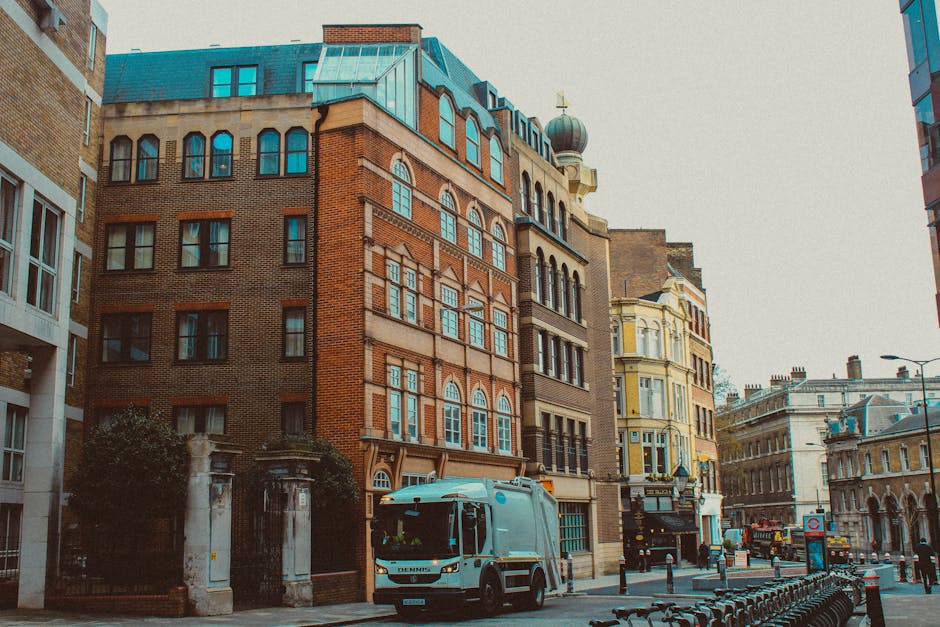 The image depicts a city street lined with multi-story buildings featuring various architectural styles, including brick and ornate facades, with some windows arched and others rectangular. In the foreground, a small waste collection truck painted in light blue is parked on the pavement, positioned beside a row of black bollards that separate the sidewalk from the street. The truck appears to be used for private waste removal, fitting within the context of independent rubbish disposal services offered by Waste Clearance Lambeth. To the left, there is a gated entrance with stone pillars and some greenery, while on the right, the street extends into the distance, showing additional vehicles and buildings. The environment is illuminated by natural daylight, and the overall scene has a typical urban atmosphere, reflecting the type of surroundings where on-site clearance and rubbish removal services might be required to manage waste outside of regular municipal collection systems.