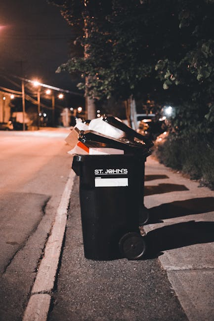 A black plastic wheeled rubbish bin labeled 'ST. JOHN'S' is positioned on a residential sidewalk beside a curb, filled with various waste items including paper and cardboard, some of which are spilling over the top. The bin is situated under a dark, leafy tree and is illuminated by nearby streetlights, creating soft shadows on the pavement. In the background, an empty street with streetlights and parked cars is visible, with the scene suggesting overnight or evening waste disposal activity. The environment appears calm and typical of an urban or suburban street where residents might leave rubbish for collection or private disposal services, such as those provided by Waste Clearance Lambeth, highlighting the importance of proper waste management practices within local communities.