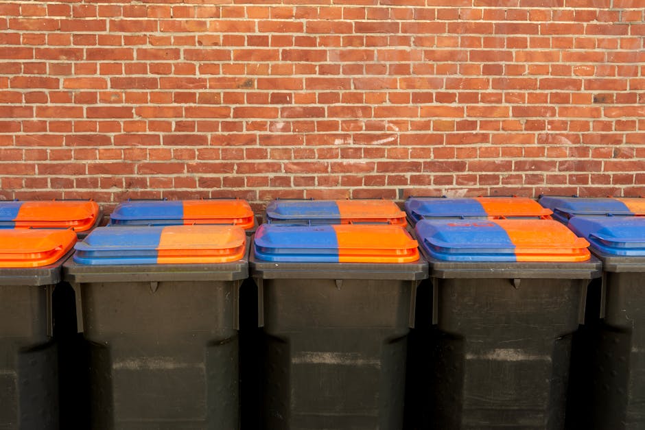 A row of identical black wheelie bins with hinged lids, arranged side by side on a paved surface in front of a red brick wall. The bins have brightly coloured lids, predominantly blue and orange, with some lids sporting a combination of these colours, indicating different waste categories or collection types. The lids are made of smooth plastic, with slight sheen reflecting ambient light, and each bin appears to be clean and uniformly maintained. The brick wall behind the bins is evenly spaced, with a rustic texture and a warm, reddish tone, providing a neutral background that highlights the bins. The scene suggests an outdoor setting, likely a commercial or residential waste disposal area, and reflects the kind of setup used in private waste collection services like Waste Clearance Lambeth, which handles rubbish removal outside of council-provided schemes. The orderly arrangement emphasizes organized waste management, typical of supplementary private disposal or on-site clearance solutions for local businesses or residents seeking alternative rubbish removal options.