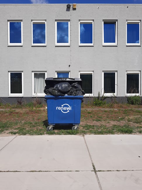 A black plastic wheeled rubbish bin labeled 'ST. JOHN'S' is positioned on a residential sidewalk beside a curb, filled with various waste items including paper and cardboard, some of which are spilling over the top. The bin is situated under a dark, leafy tree and is illuminated by nearby streetlights, creating soft shadows on the pavement. In the background, an empty street with streetlights and parked cars is visible, with the scene suggesting overnight or evening waste disposal activity. The environment appears calm and typical of an urban or suburban street where residents might leave rubbish for collection or private disposal services, such as those provided by Waste Clearance Lambeth, highlighting the importance of proper waste management practices within local communities.