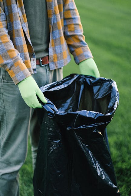 A person wearing a grey shirt and a yellow and grey checkered jacket, along with bright green disposable gloves, stands outdoors in a grassy area. They are holding a large, open black plastic rubbish bag with both hands, preparing for waste collection or disposal. The background features a lush green lawn, indicating a clean and maintained environment suitable for waste clearance activities. The image emphasizes the hands-on approach of private rubbish removal services, such as those offered by Waste Clearance Lambeth, demonstrating a professional and environmentally conscious method of waste management. The focus on the gloves and bag highlights the importance of proper handling and disposal of rubbish, aligning with the context of independent collection or on-site clearance practices, frequently used as alternative waste handling options outside of traditional council refuse collection systems.