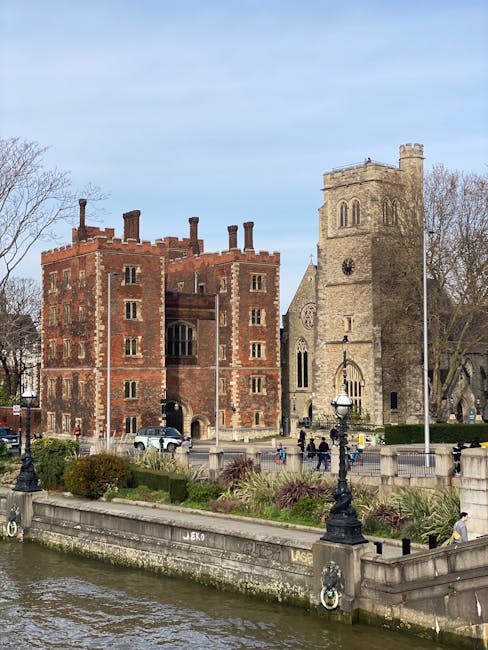 A historic brick and stone building situated beside a river, with a red brick section featuring crenellated parapets and multiple small window openings, alongside a taller grey stone tower with arched windows and a clock face near the top. The scene includes a paved promenade with decorative black lampposts, some greenery, and a low stone wall bordering the riverbank. Several pedestrians are visible walking along the promenade, and a few vehicles are parked near the building. The background shows leafless trees and a partly cloudy sky, suggesting a cool season. This setting aligns with private or alternative waste management contexts, where rubbish removal services by Waste Clearance Lambeth might be utilized for clearing debris around historic sites or along riverside areas, ensuring preservation and cleanliness without reliance on local authority disposal.