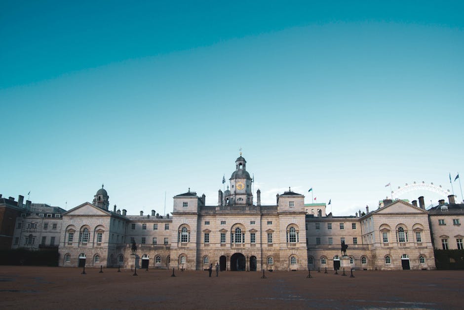 A large historic building with a symmetrical façade constructed from light-colored stone, featuring multiple arched and rectangular windows, decorative cornices, and a central clock tower topped with a dome and spire. The structure is situated on a spacious paved area with a few small, dark bollards aligned in front, and several small figures of people walking or standing near the entrance. The background includes a clear, bright blue sky. The scene is outdoors, and the building's appearance and surrounding open space suggest it could be an official or civic building, with no visible rubbish or waste materials present. This setting relates to options for private or independent waste collection for properties like this, in a context supported by waste clearance services such as Waste Clearance Lambeth.