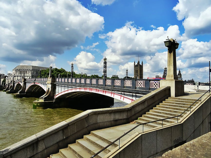 A view of Westminster Bridge spanning the River Thames in London, with the bridge's ornate metal railings painted in white and pink hues visible in the foreground. The stone staircase descending from the riverside promenade is seen on the right side of the image, equipped with metal handrails along the steps. The river below has a murky greenish-brown tone, with some moss or algae growth visible on the submerged sides of the bridge supports. In the background, prominent landmarks including the gothic-style clock tower of Westminster, known as Big Ben, and the Palace of Westminster with its pointed spires are visible beneath a partly cloudy sky with white, billowing clouds set against a blue sky. The scene is well-lit by natural daylight, creating a clear and detailed urban landscape that reflects the historic and architectural elements of central London. This environmental setting relates to urban infrastructure, which can be associated with the context of city waste management, possibly highlighting considerations for public space maintenance or alternative disposal methods in busy city centers, as handled by waste clearance services like Waste Clearance Lambeth.