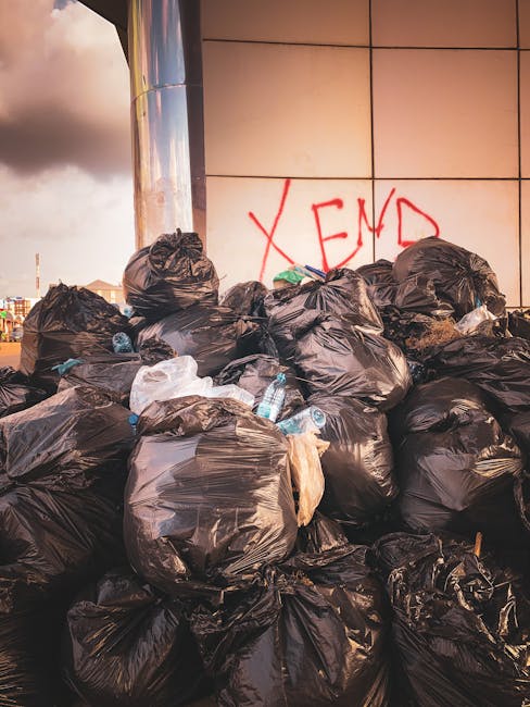 A large pile of black garbage bags, some torn or partially open, stacked against a tiled wall outdoors. The bags appear to contain mixed waste, with some plastic bottles and packaging visibly protruding from openings. Behind the pile, the tiled wall features red spray-painted graffiti with the word 'XEND'. The setting is urban, likely an alley or backstreet, with a cloudy sky seen in the background. The scene exemplifies the type of waste accumulation that waste clearance services, such as Waste Clearance Lambeth, might manage through on-site collection or private waste handling solutions. The lighting is natural, possibly during late afternoon, highlighting the textures of the tightly packed plastic bags and the smooth, reflective surface of the tiled wall. This image illustrates an example of unmanaged rubbish accumulation requiring professional removal to prevent environmental and health hazards typical of urban waste storage areas.
