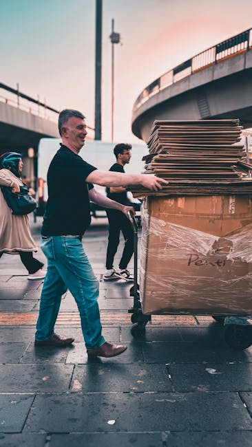 A man with short dark hair, wearing a black T-shirt, blue jeans, and brown shoes, is standing outdoors under a large road bridge during an evening or late afternoon, with the sky displaying a warm, pinkish hue. He is pushing a large, heavy-duty wheeled bin filled with flattened cardboard boxes, some with visible text and logos, which are stacked neatly and securely inside the bin. Behind him, a young man with dark hair and glasses, dressed in a black jacket and dark pants, stands nearby, while a woman in a beige coat and backpack is walking past on the left side of the image. The setting appears to be a busy urban area with paved street surfaces, and the background shows the underside of an overpass with metal railings and supporting columns, indicating a location suitable for private waste collection or alternative rubbish removal services like those provided by Waste Clearance Lambeth, in an environment where on-site waste handling is occurring.