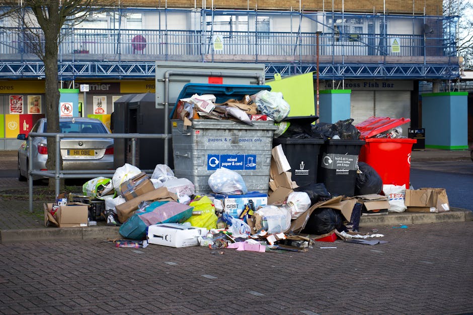 A large collection of mixed household waste and recyclable materials is piled around a central grey recycling bin on a paved urban parking area. The bin, labeled for mixed paper and cardboard, is filled to the brim with various papers, boxes, and packaging, some of which are overflowing onto the surrounding ground. Adjacent to the grey bin are several black and red waste bags and closed cardboard boxes, containing additional rubbish. Behind the waste containers, there is a parking space where a grey hatchback car with a partially visible number plate is parked. In the background, a commercial building with a blue scaffolding structure and signage is visible, indicating a commercial or retail area. A metal railing runs along the edge of the pavement, partially separating the waste area from the parked vehicle and pedestrian space. The scene is captured in daylight, with natural light illuminating the cluttered environment, reflecting situations where private waste collection services like Waste Clearance Lambeth might be involved in on-site rubbish removal or alternative waste handling beyond standard council collection. The environment and visible waste items suggest an effort to manage rubbish independently, aligning with private clearance services.