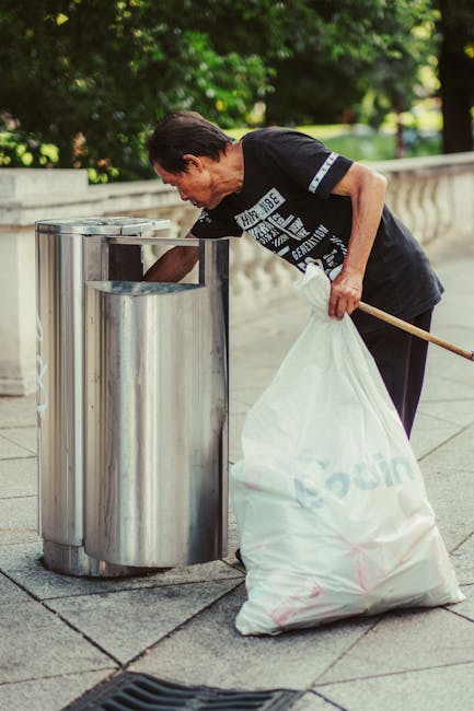 A middle-aged man with dark hair wearing a black T-shirt with white text is seen disposing of rubbish into a modern, stainless steel public litter bin on a paved sidewalk. He leans forward slightly, holding a large white plastic bag filled with waste in his left hand while using a wooden litter picker in his right hand to guide debris into the bin's opening. The bin is cylindrical with a smooth, reflective surface, positioned on a paved surface with visible lines and a slight drainage grate nearby. Behind him, there is a low stone or concrete balustrade and a backdrop of lush green trees, indicating an outdoor urban or park setting. The scene is well-lit with natural daylight, illustrating a typical effort of independent waste collection or rubbish disposal by a private individual or service, consistent with alternative waste handling practices often seen in rubbish removal or street cleaning scenarios. Waste Clearance Lambeth and similar services might be called upon for larger or more complex clearance needs, but this image depicts straightforward rubbish disposal in a public environment.